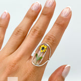 A silver ring with a clear oval-shaped setting containing a pressed yellow sunflower. The ring is designed with a curved adjustable band.