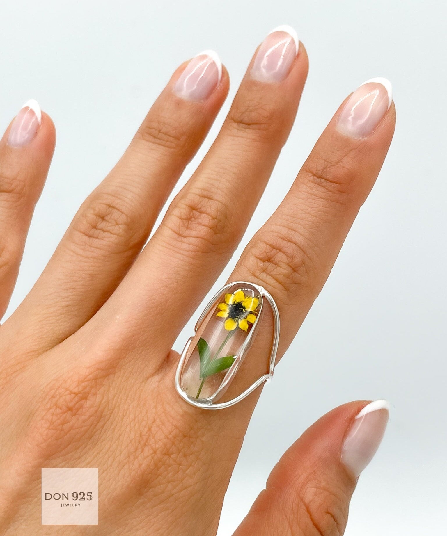 A silver ring with a clear oval-shaped setting containing a pressed yellow sunflower. The ring is designed with a curved adjustable band.