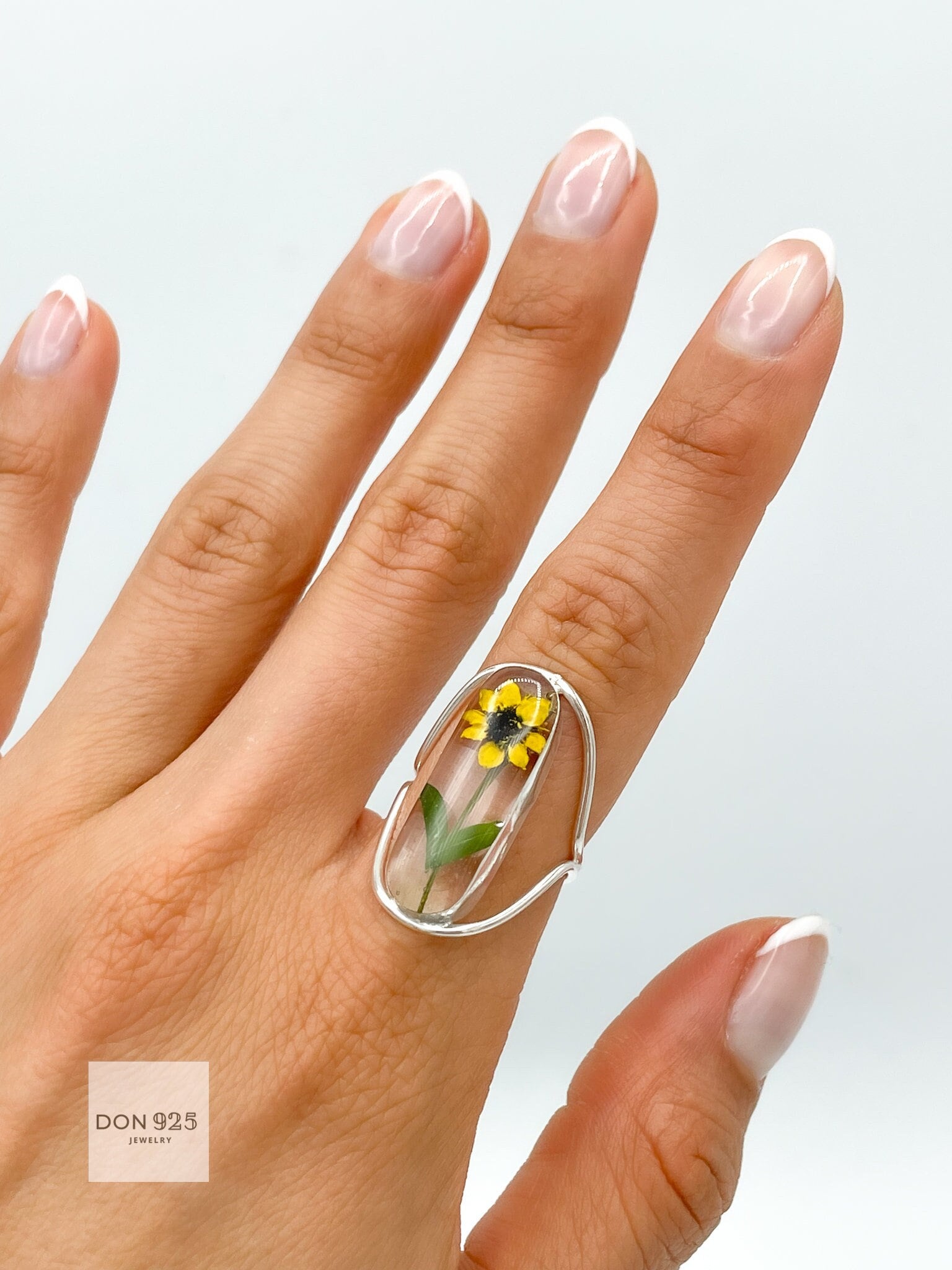 A silver ring with a clear oval-shaped setting containing a pressed yellow sunflower. The ring is designed with a curved adjustable band.