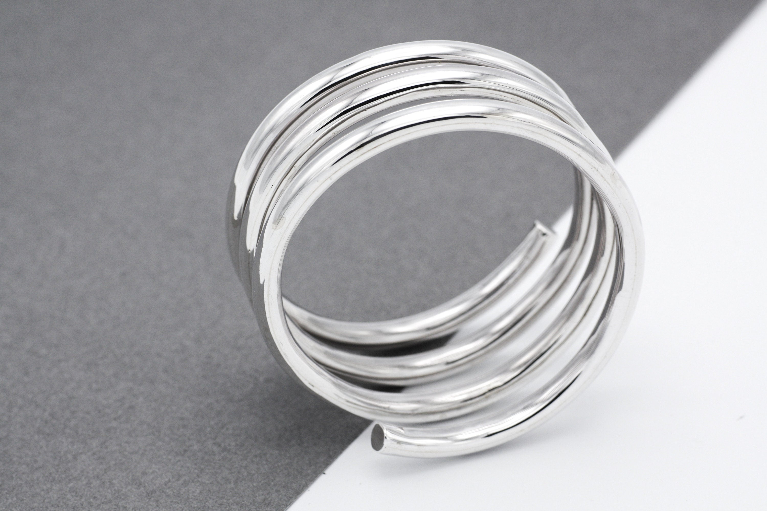 May include: A silver-colored ring with a spiral design. The ring is composed of multiple, tightly wound bands, creating a layered effect. The bands have a smooth, reflective surface, and the ring's design is modern and minimalist, suitable for everyday wear.