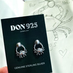 A pair of sterling silver earrings in the shape of a crab, holding a gray pearl