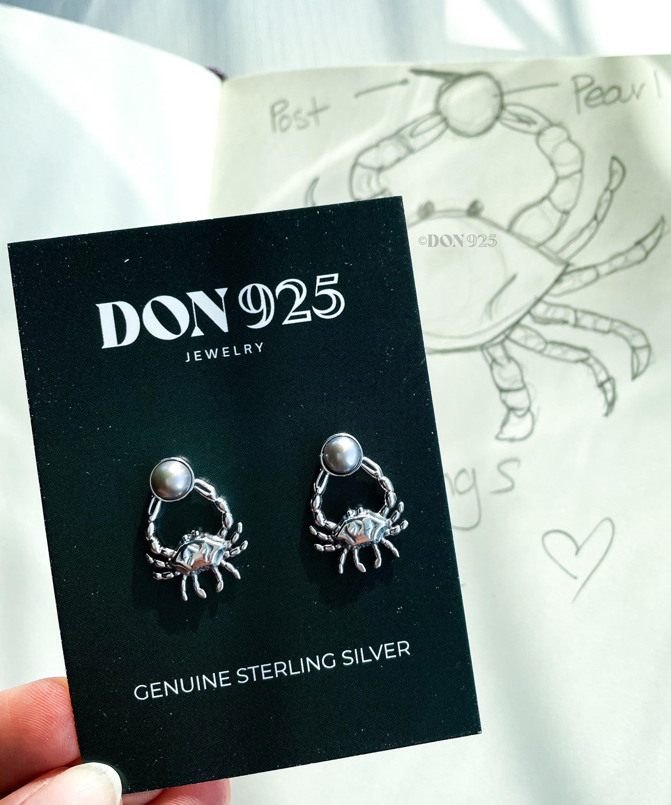 A pair of sterling silver earrings in the shape of a crab, holding a gray pearl
