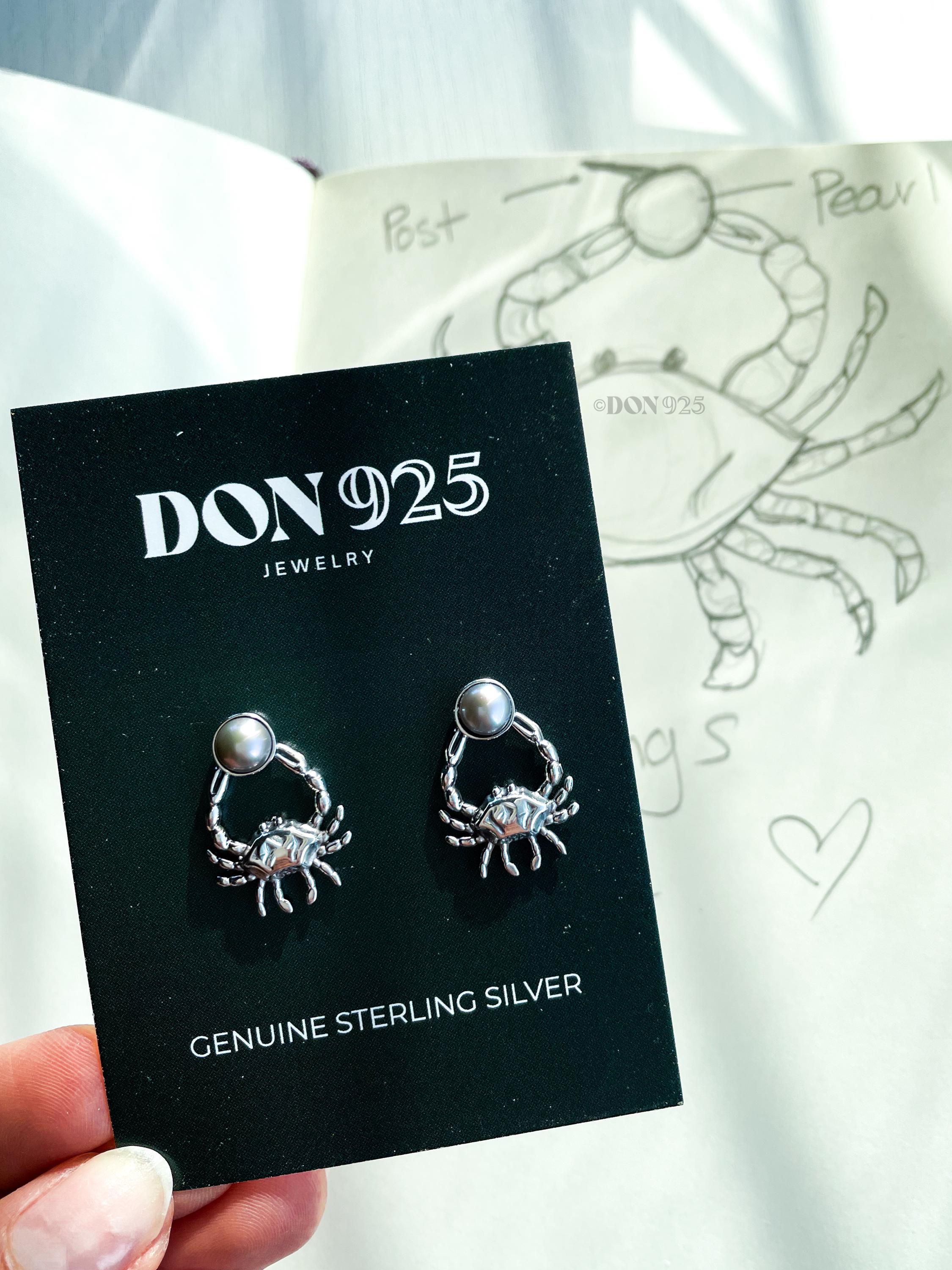 A pair of sterling silver earrings in the shape of a crab, holding a gray pearl