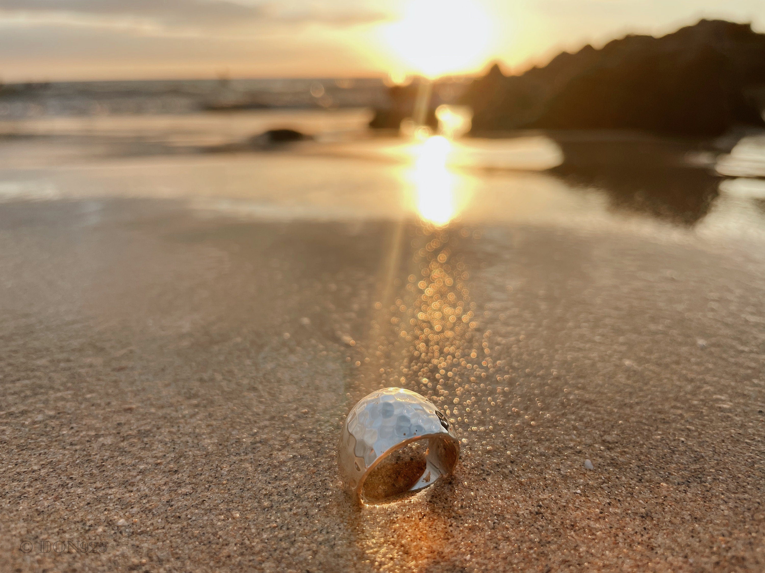May include: A silver hammered ring on a sandy beach with the sun setting in the background.
