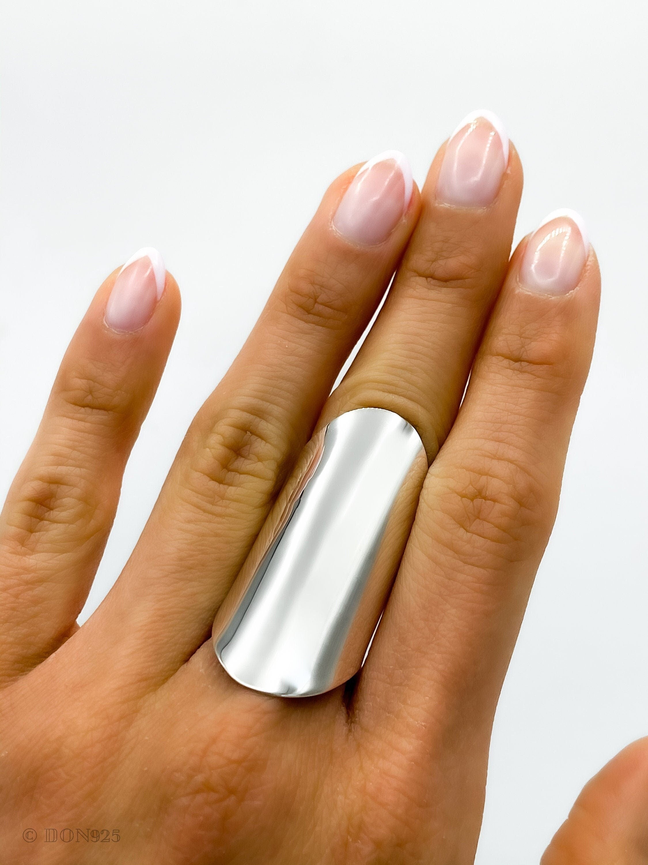 May include: A silver, wide, flat band ring worn on the middle finger of a hand with white nail polish.