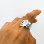 May include: A silver hammered ring with a wide band. The ring is worn on a person's finger.