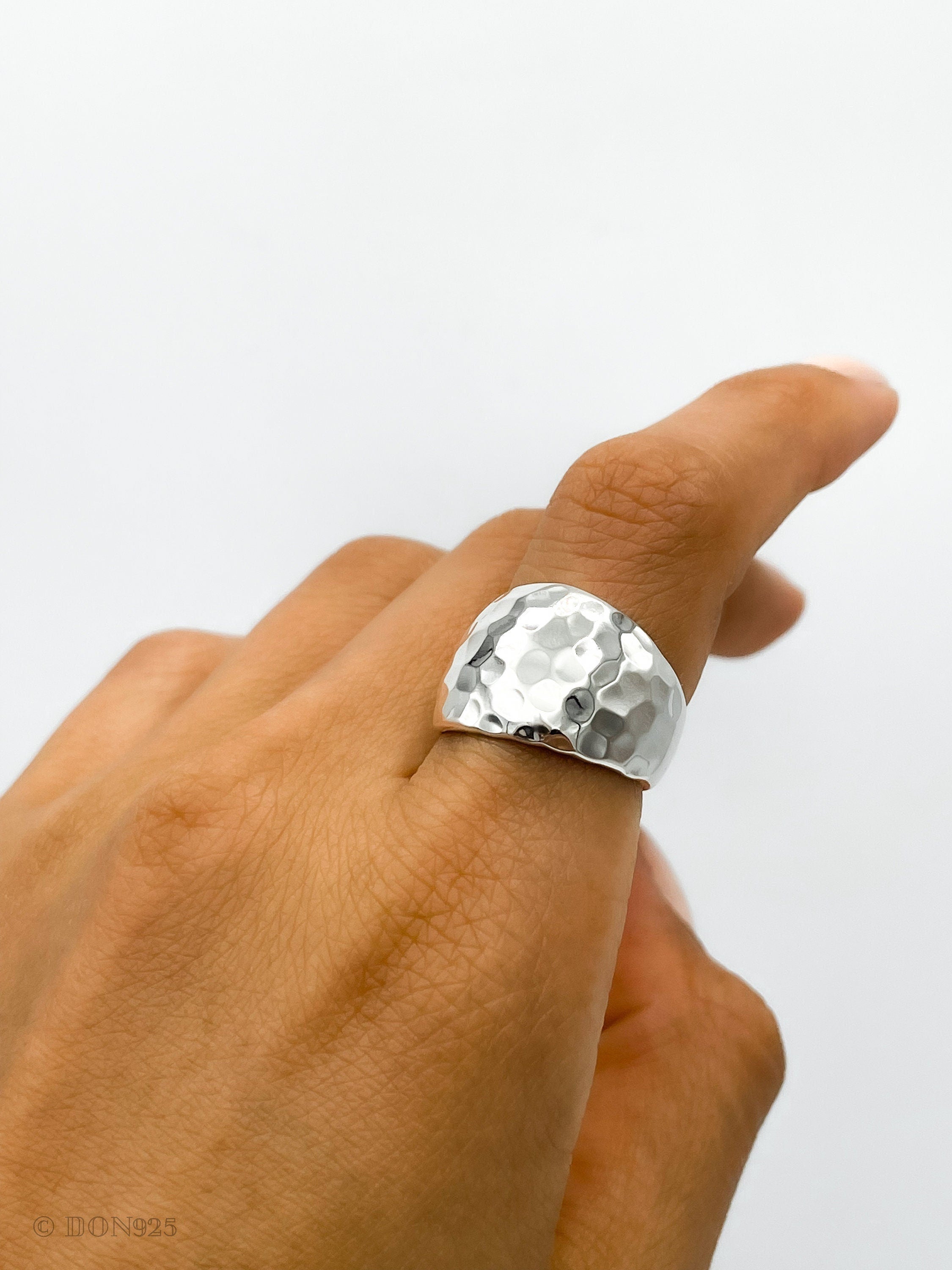May include: A silver hammered ring with a wide band. The ring is worn on a person's finger.