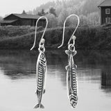 May include: Silver fish-shaped earrings with intricate detailing, suspended from curved ear wires. The earrings are displayed against a backdrop of a serene lake and a mountainous landscape. The jewelry is from the brand "DON 925 JEWELRY."