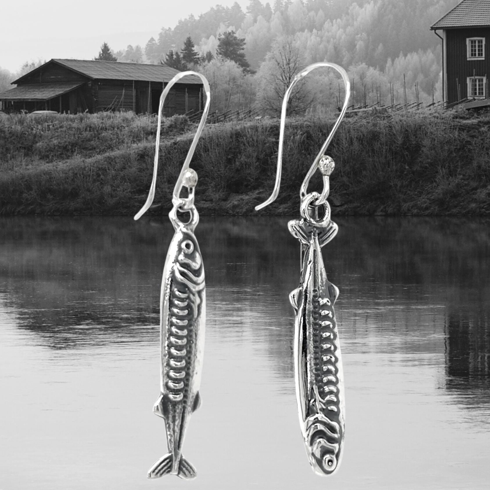 May include: Silver fish-shaped earrings with intricate detailing, suspended from curved ear wires. The earrings are displayed against a backdrop of a serene lake and a mountainous landscape. The jewelry is from the brand "DON 925 JEWELRY."
