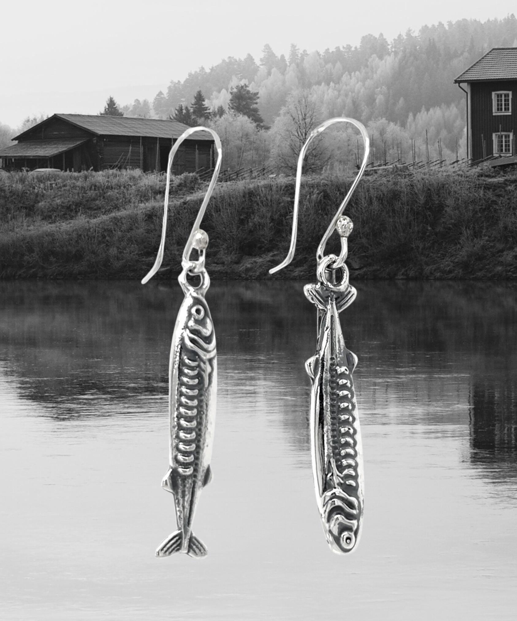 May include: Silver fish-shaped earrings with intricate detailing, suspended from curved ear wires. The earrings are displayed against a backdrop of a serene lake and a mountainous landscape. The jewelry is from the brand "DON 925 JEWELRY."