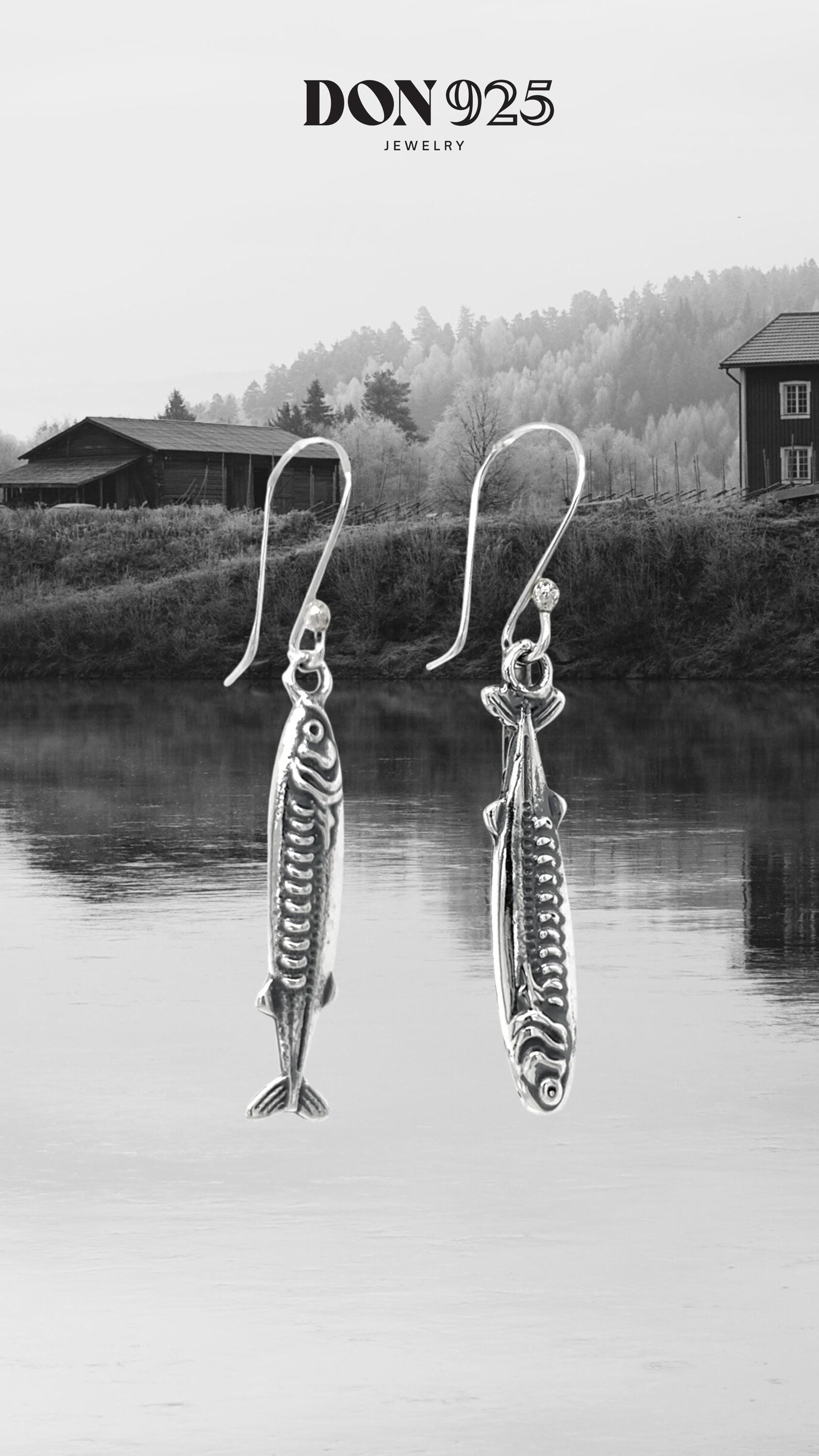 May include: Silver fish-shaped earrings with intricate detailing, suspended from curved ear wires. The earrings are displayed against a backdrop of a serene lake and a mountainous landscape. The jewelry is from the brand "DON 925 JEWELRY."