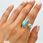May include: A silver ring with two turquoise stones. The larger stone is a crescent shape and the smaller stone is round. The ring is on a person's finger.