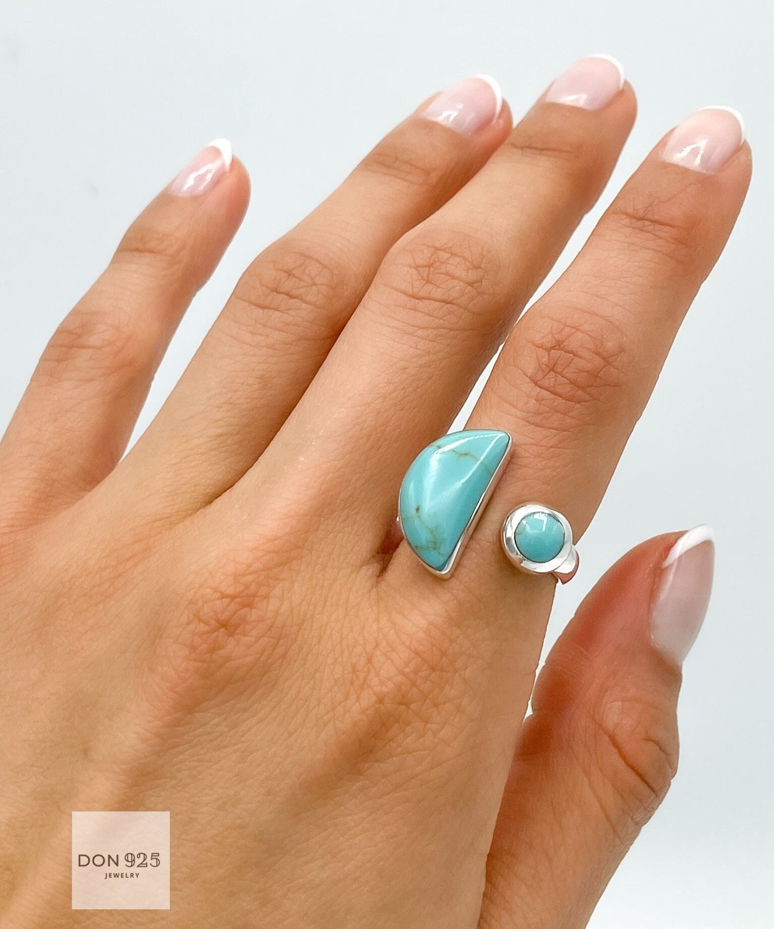 May include: A silver ring with two turquoise stones. The larger stone is a crescent shape and the smaller stone is round. The ring is on a person's finger.