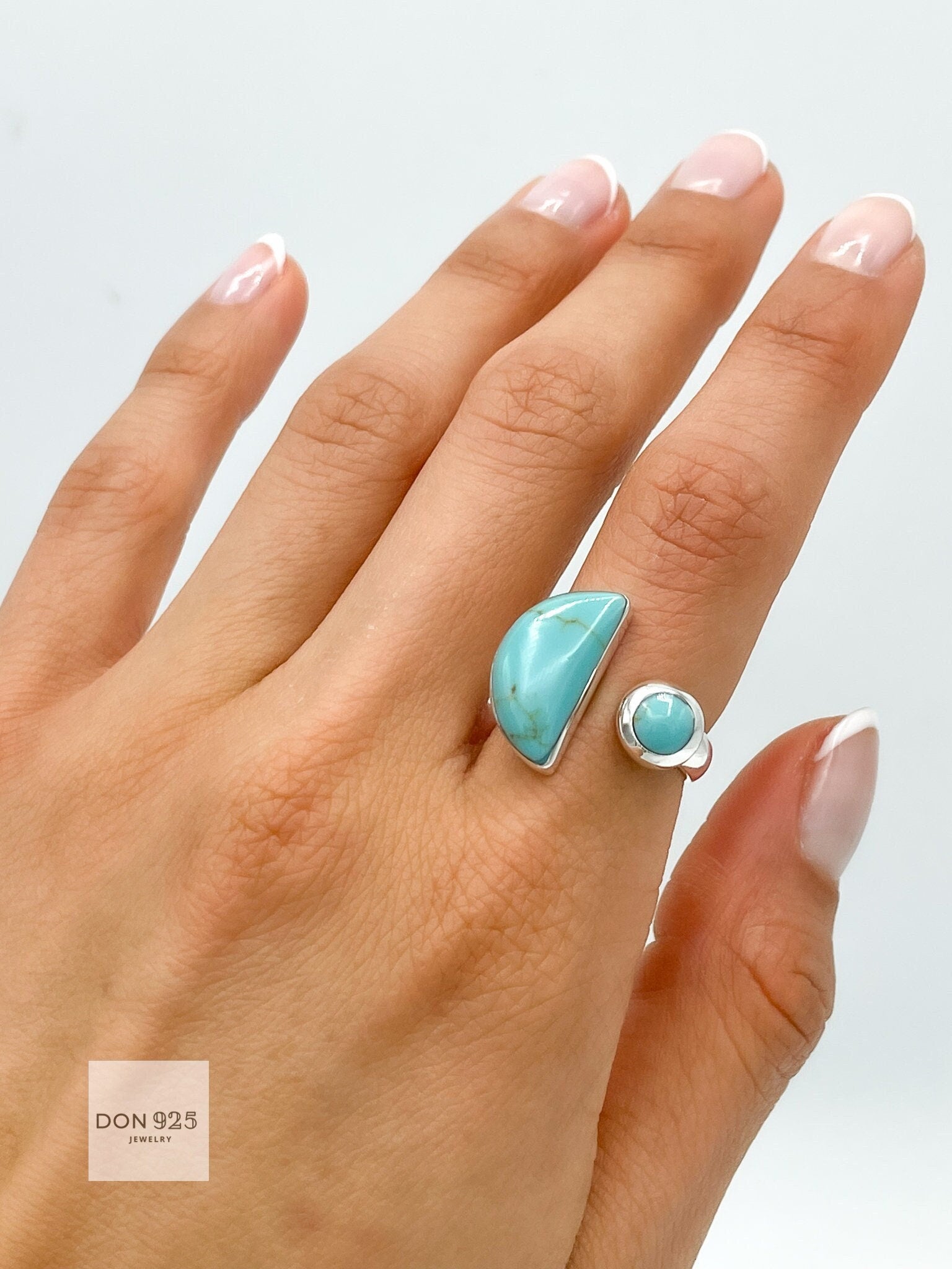May include: A silver ring with two turquoise stones. The larger stone is a crescent shape and the smaller stone is round. The ring is on a person's finger.