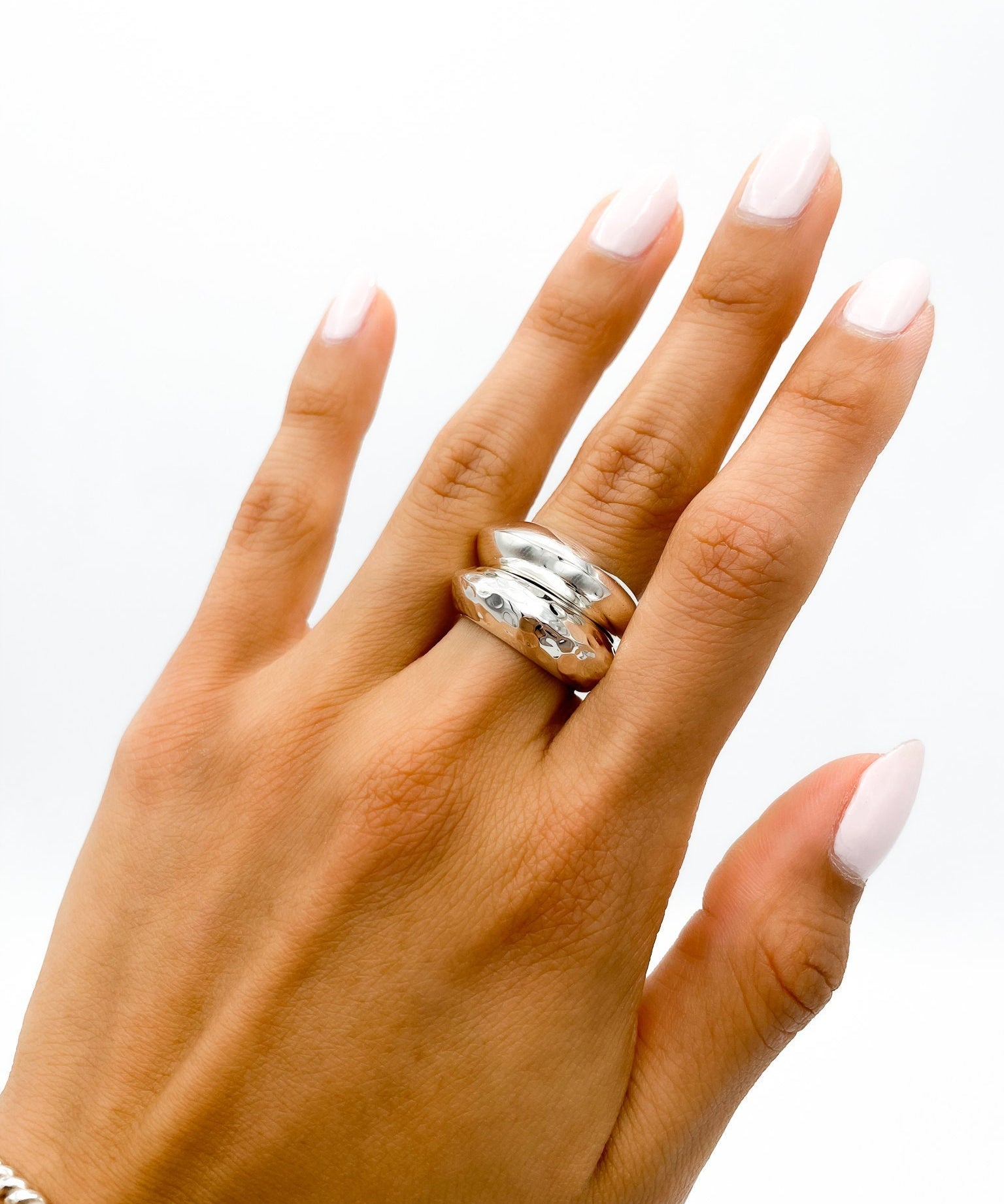 May include: A silver ring with a hammered texture. The ring is worn on the middle finger of a hand with light pink nail polish.