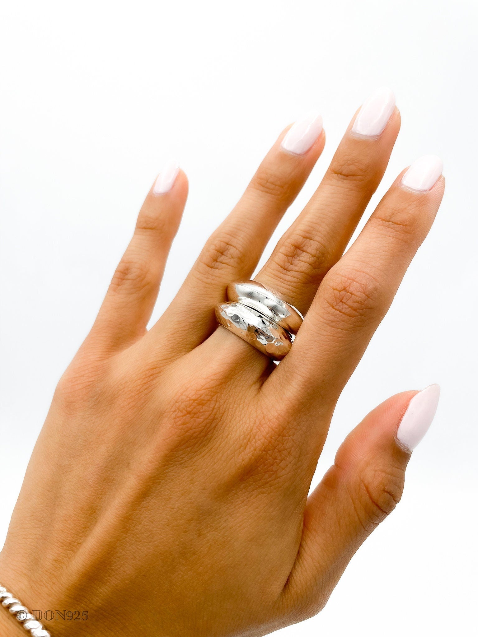 May include: A silver ring with a hammered texture. The ring is worn on the middle finger of a hand with light pink nail polish.