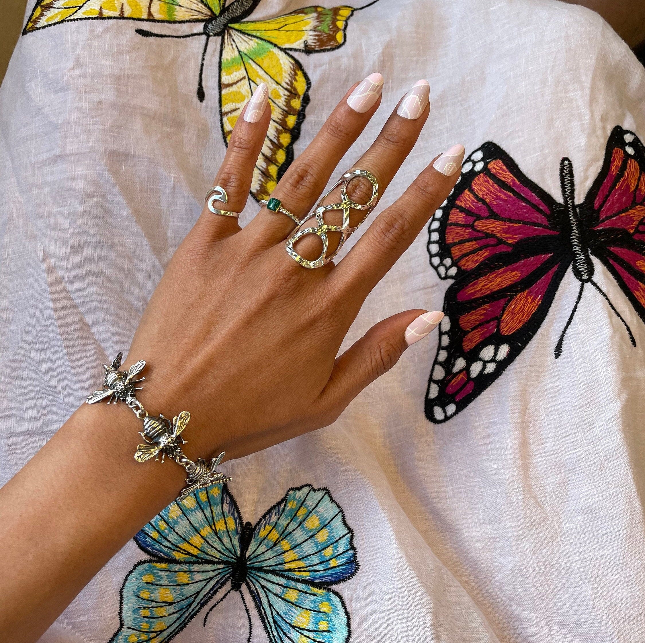 May include: A silver bracelet with a bee design. The bracelet is on a person's wrist. The person is wearing several silver rings on their fingers, including a ring with a green stone.