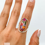 May include: A silver ring with a clear oval-shaped stone containing pressed flowers. The flowers are a variety of colors, including pink, red, orange, yellow, and blue.