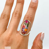 May include: A silver ring with a clear oval-shaped stone containing pressed flowers. The flowers are a variety of colors, including pink, red, orange, yellow, and blue.