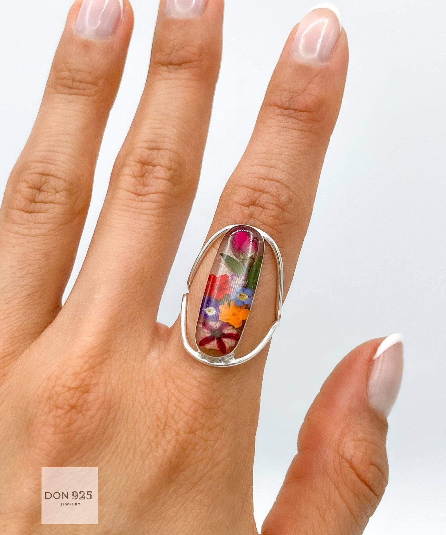 May include: A silver ring with a clear oval-shaped stone containing pressed flowers. The flowers are a variety of colors, including pink, red, orange, yellow, and blue.