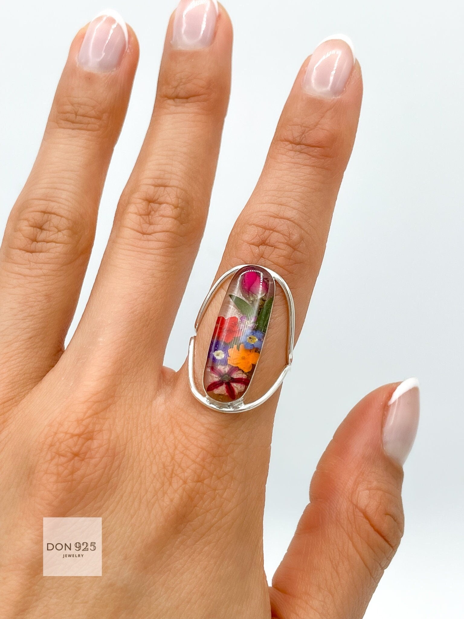 May include: A silver ring with a clear oval-shaped stone containing pressed flowers. The flowers are a variety of colors, including pink, red, orange, yellow, and blue.