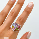 May include: A silver heart-shaped ring with a clear resin inlay featuring pressed flowers in shades of pink, purple, blue, and yellow. The ring is worn on a finger with a white French manicure.
