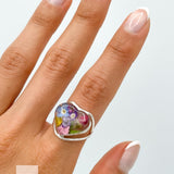 May include: A silver heart-shaped ring with a clear resin inlay featuring pressed flowers in shades of pink, purple, blue, and yellow. The ring is worn on a finger with a white French manicure.
