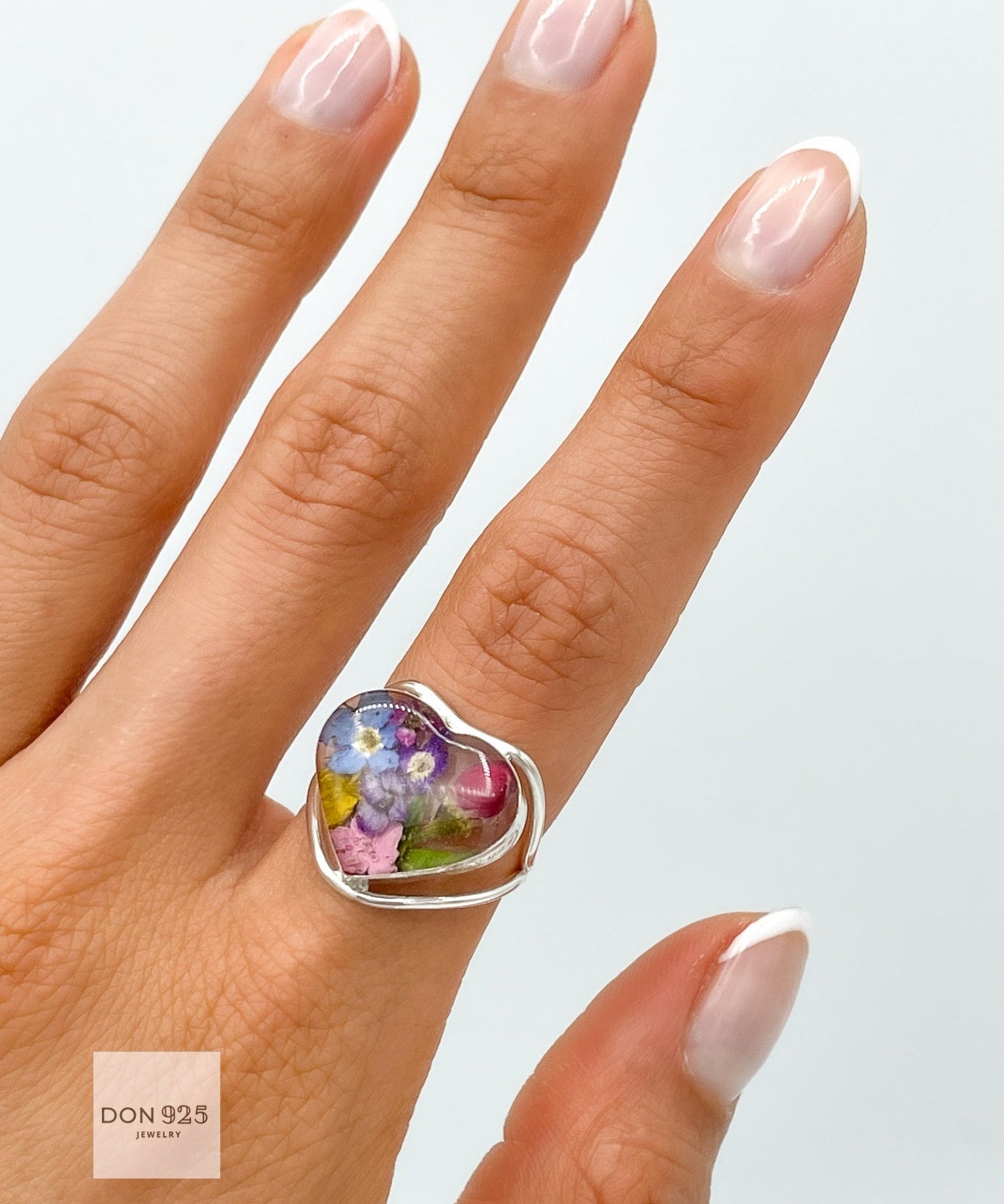May include: A silver heart-shaped ring with a clear resin inlay featuring pressed flowers in shades of pink, purple, blue, and yellow. The ring is worn on a finger with a white French manicure.