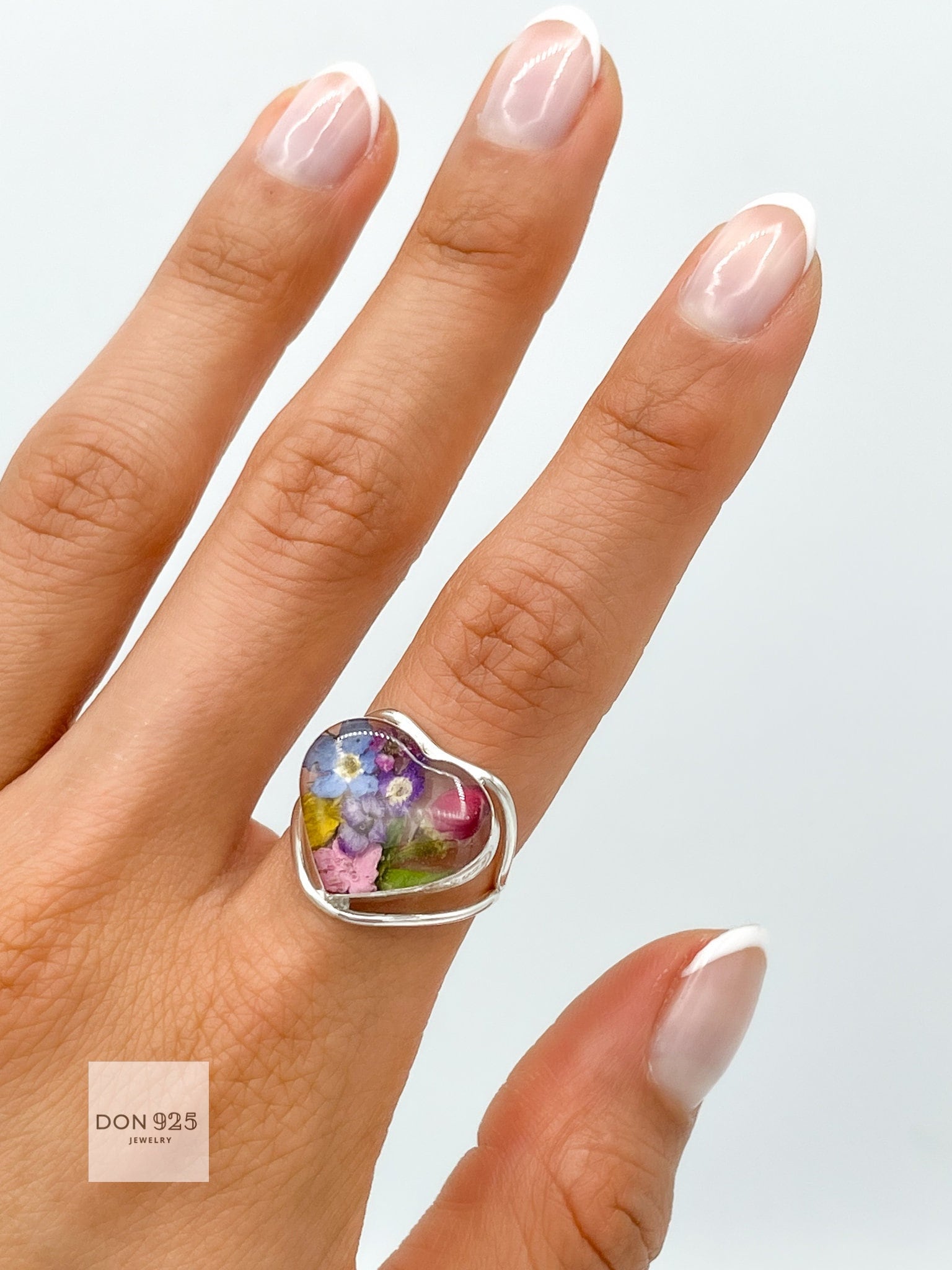 May include: A silver heart-shaped ring with a clear resin inlay featuring pressed flowers in shades of pink, purple, blue, and yellow. The ring is worn on a finger with a white French manicure.