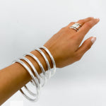 May include: A set of six silver bangles stacked on a person's wrist. The bangles are smooth and shiny, and they are all the same width. The bangles are a popular jewelry item for women.