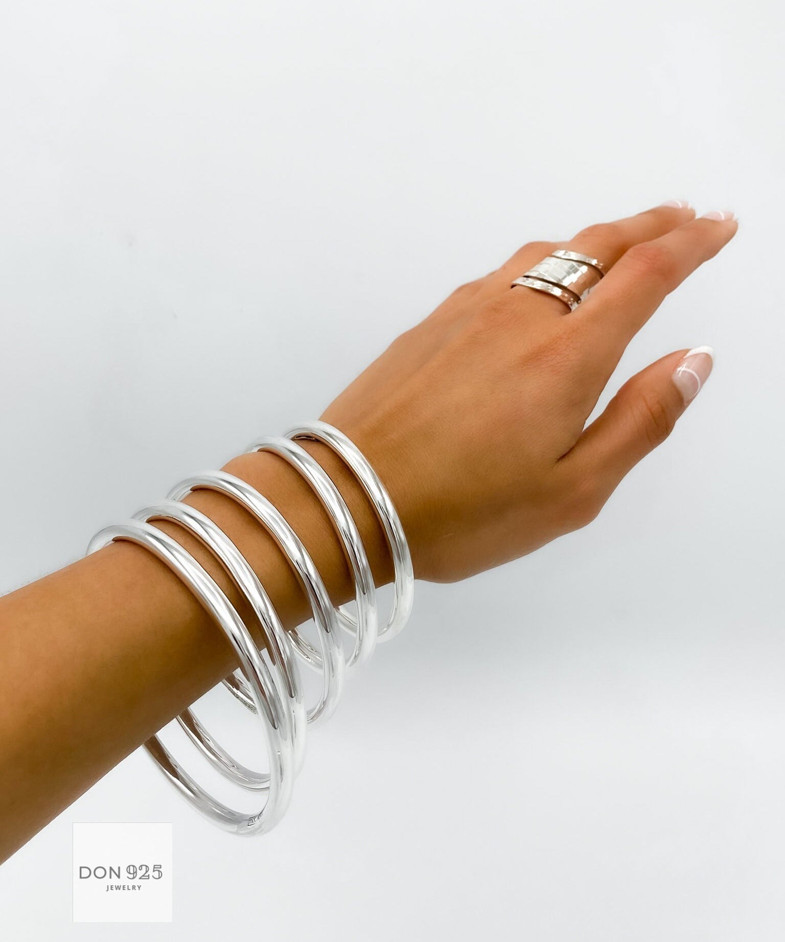 May include: A set of six silver bangles stacked on a person's wrist. The bangles are smooth and shiny, and they are all the same width. The bangles are a popular jewelry item for women.