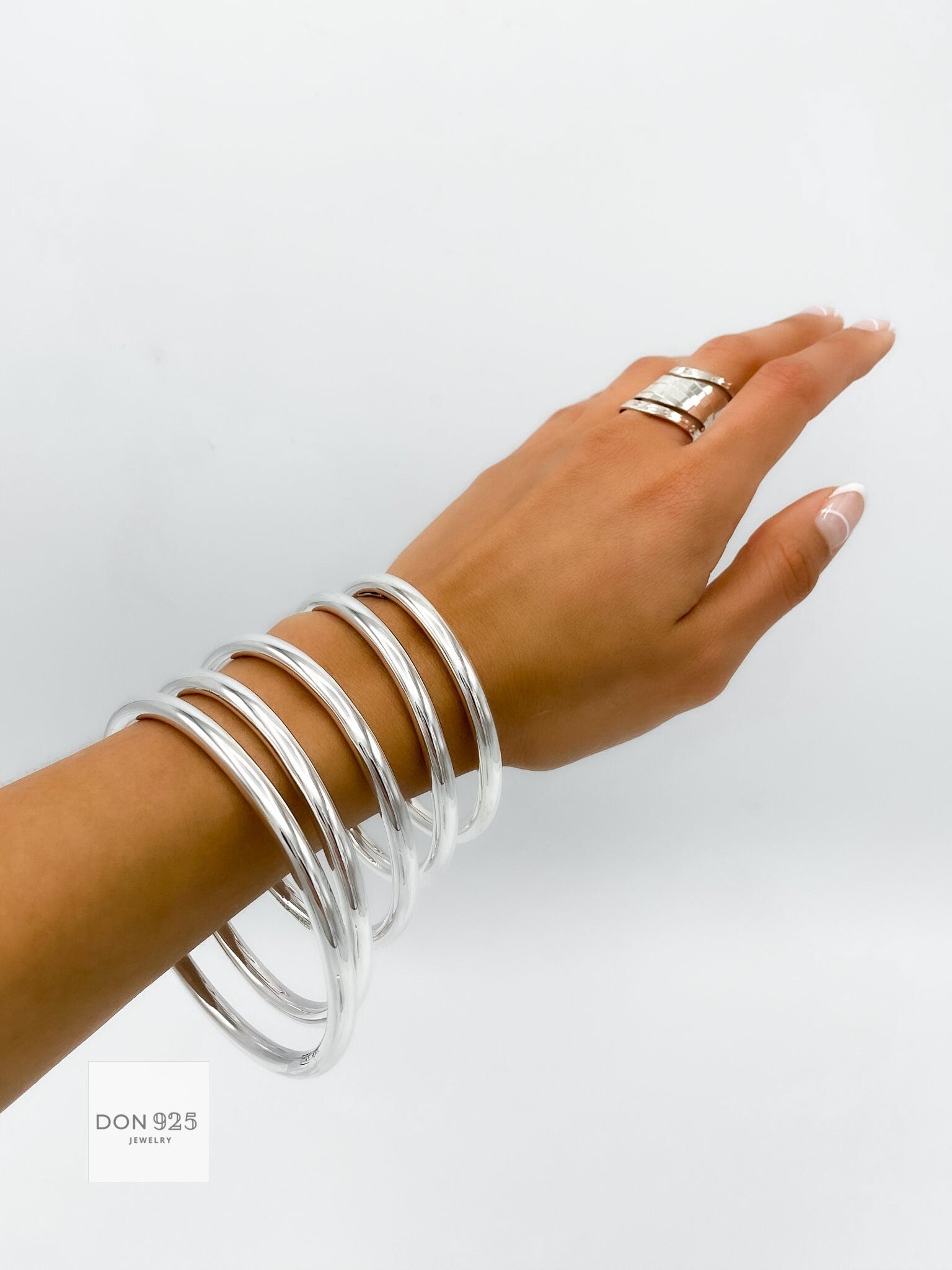 May include: A set of six silver bangles stacked on a person's wrist. The bangles are smooth and shiny, and they are all the same width. The bangles are a popular jewelry item for women.