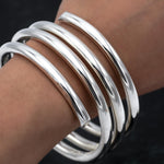 May include: A set of five shiny, silver-colored bangles. The bracelets are thick and rounded, with a smooth, reflective surface. The bangles are stacked on a wrist, showcasing their simple, elegant design. The jewelry is a classic accessory.