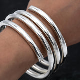 May include: A set of five shiny, silver-colored bangles. The bracelets are thick and rounded, with a smooth, reflective surface. The bangles are stacked on a wrist, showcasing their simple, elegant design. The jewelry is a classic accessory.