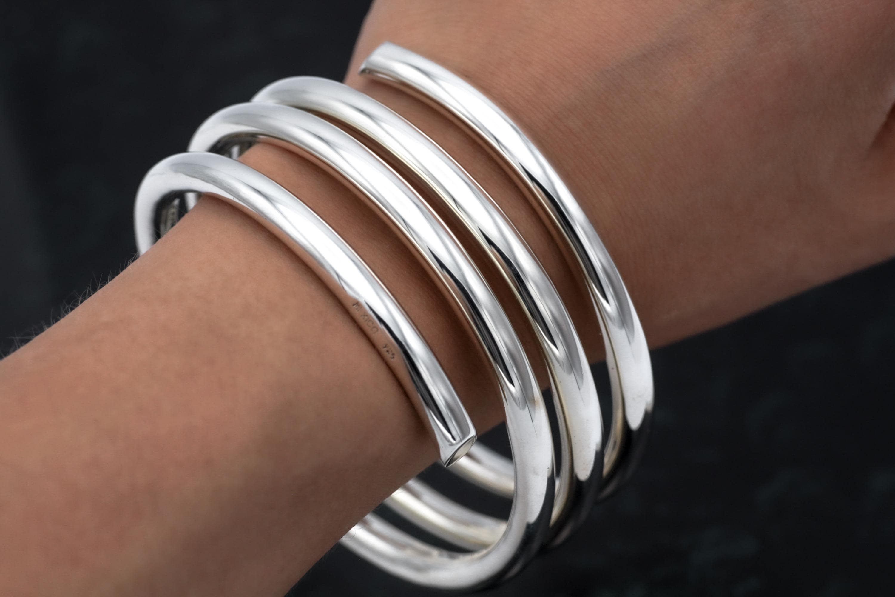 May include: A set of five shiny, silver-colored bangles. The bracelets are thick and rounded, with a smooth, reflective surface. The bangles are stacked on a wrist, showcasing their simple, elegant design. The jewelry is a classic accessory.
