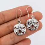 May include: Silver dangle earrings featuring a textured, round sand dollar design with a starfish in the center.