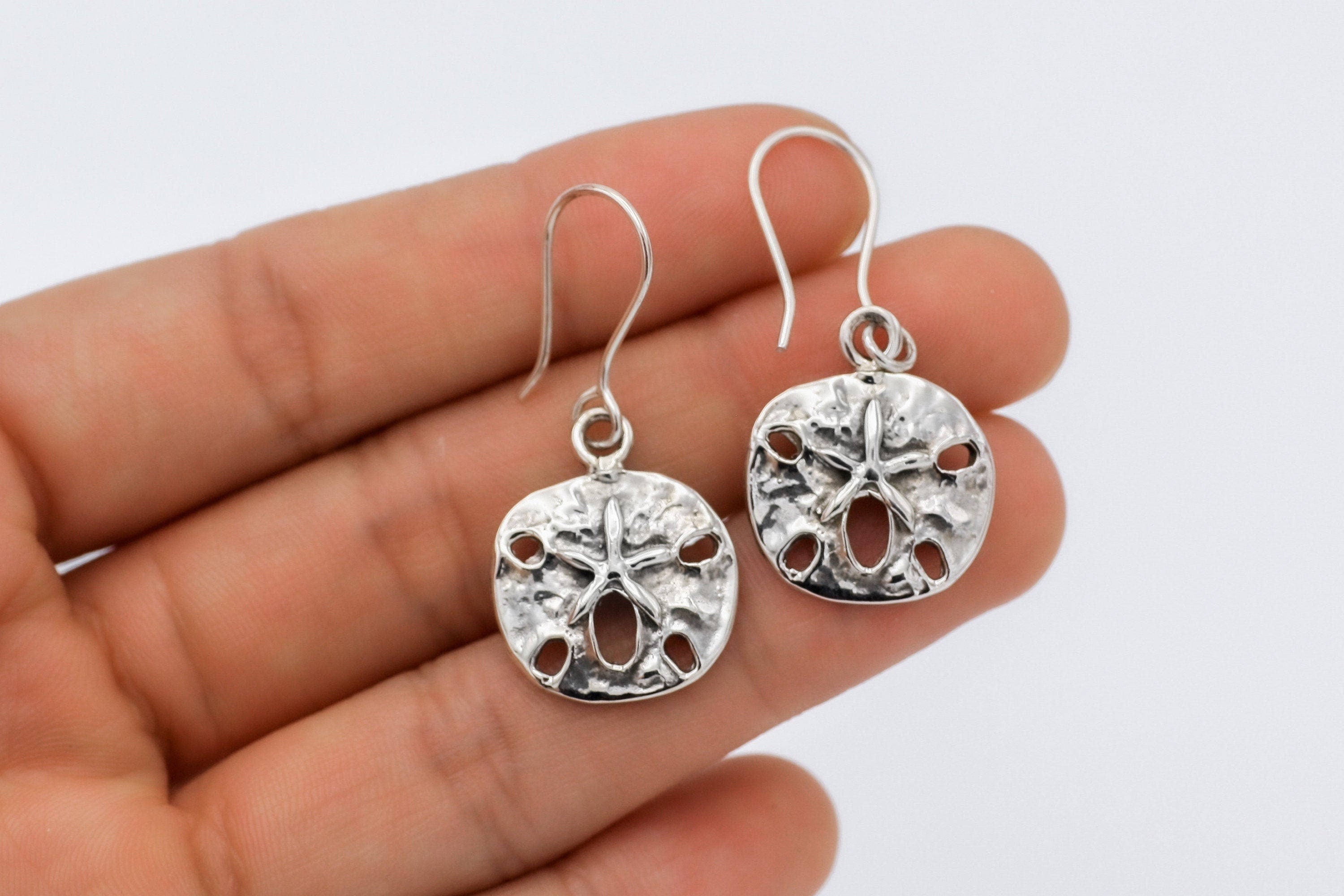 May include: Silver dangle earrings featuring a textured, round sand dollar design with a starfish in the center.