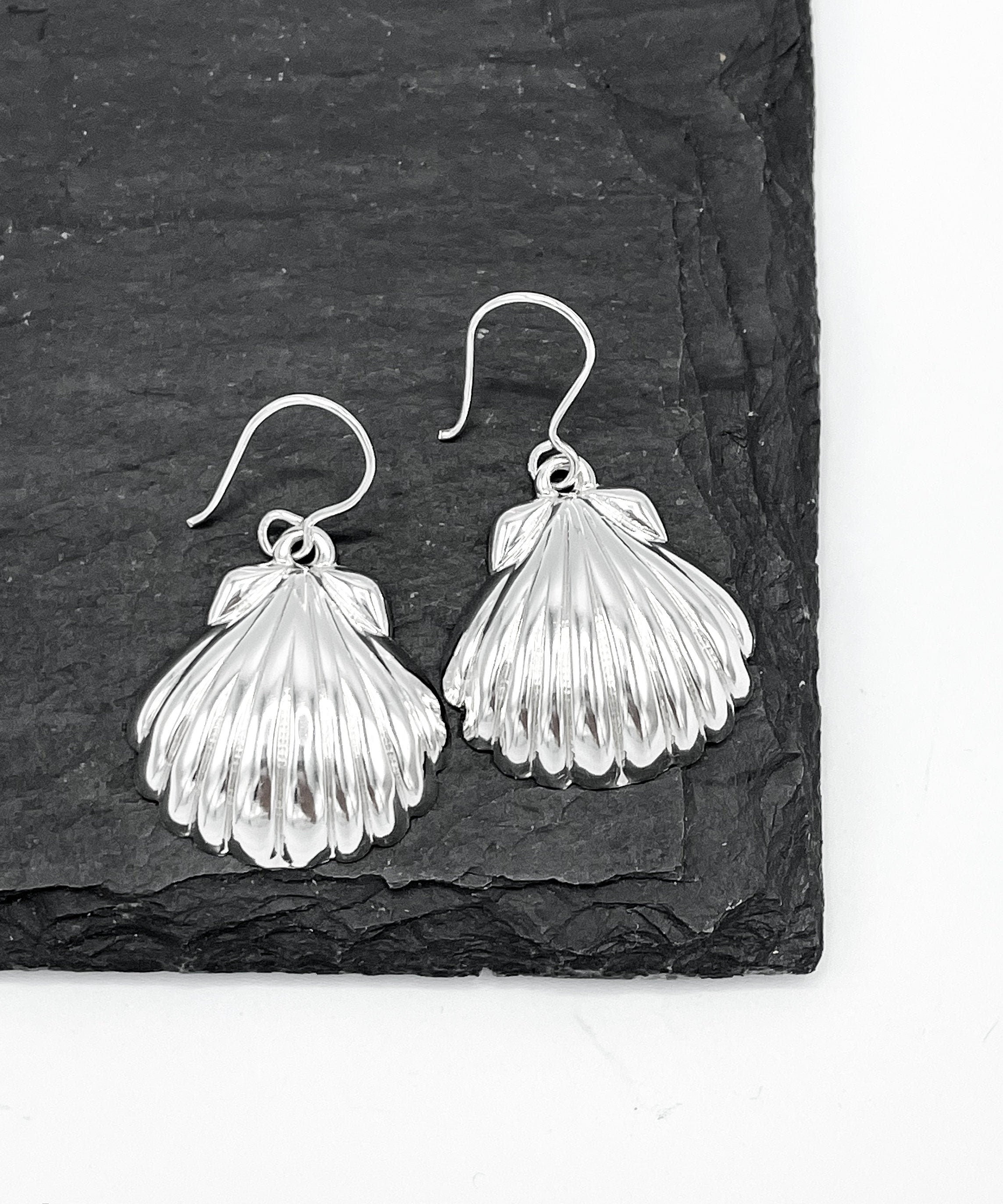 May include: A pair of silver earrings shaped like seashells. The earrings are hanging from a black slate surface.