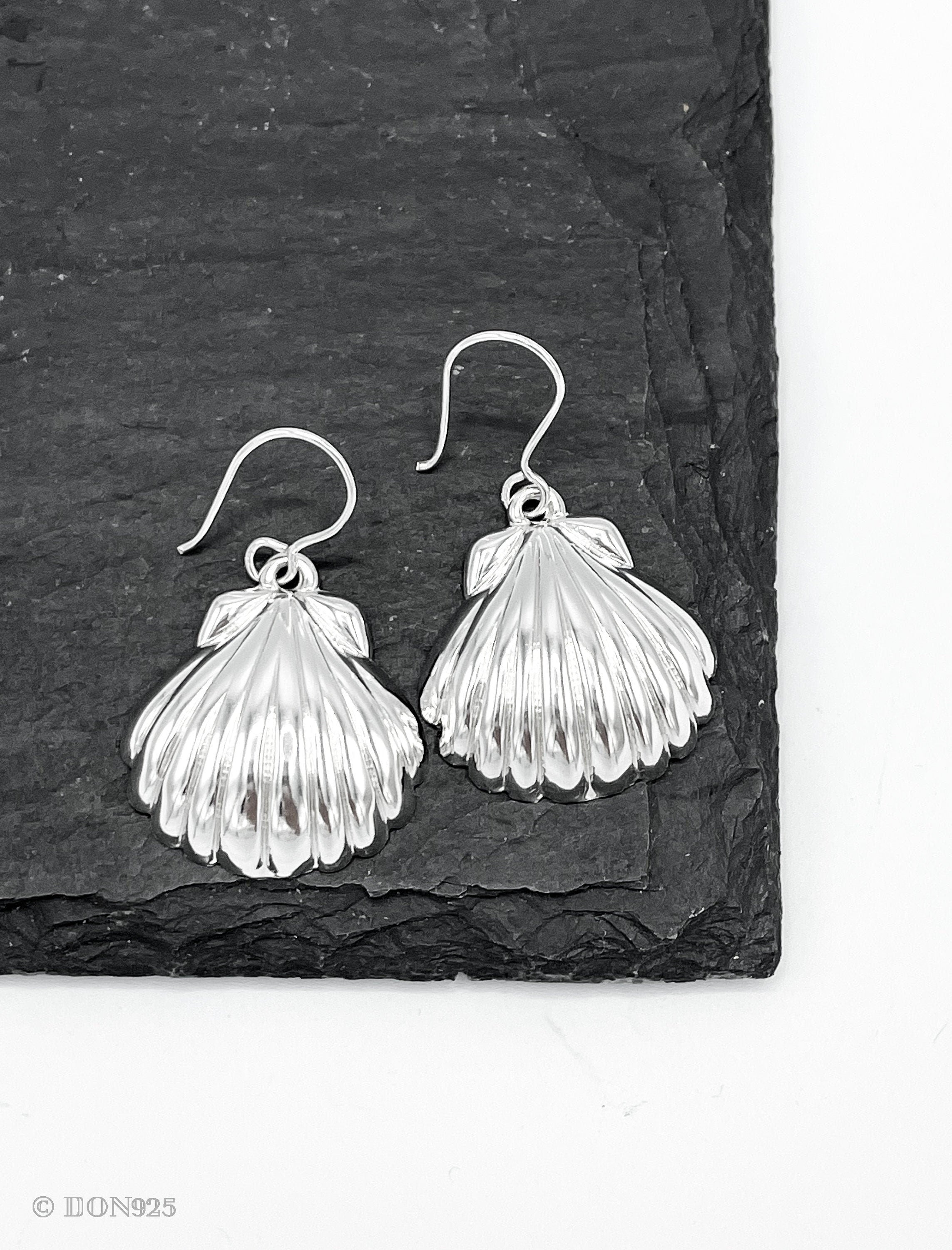 May include: A pair of silver earrings shaped like seashells. The earrings are hanging from a black slate surface.